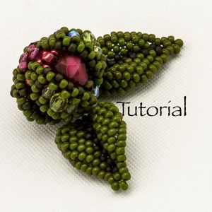 May include: A beaded flower with green leaves. The flower is made with small, round beads in shades of green, brown, and red. The center of the flower has a large, red, faceted bead. The leaves are made with green beads and have a pointed shape. The word "Tutorial" is written in black text below the flower.