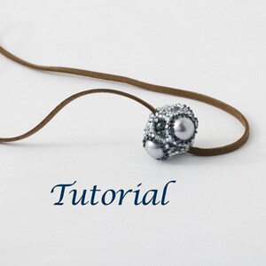 May include: A silver and white beaded necklace with a pendant featuring three white pearls. The necklace is on a brown leather cord. The word "Tutorial" is written in blue below the necklace.
