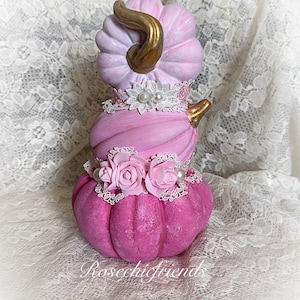 May include: A stacked pumpkin decoration featuring three tiers in shades of light pink to deep magenta. The top pumpkin has a gold stem, while the middle and bottom sections are adorned with white lace trim, faux pearls, and pink fabric roses.