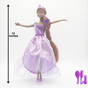May include: A doll, approximately 12 inches tall, wearing a lavender dress with a white floral pattern and a sheer overlay. The doll has long brown hair and is accessorized with a crown and a sash. A set of purple hair accessories is included.