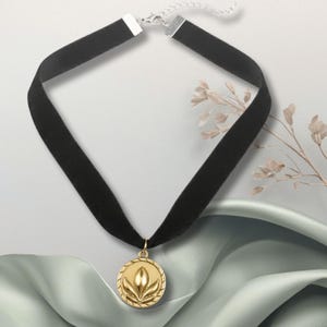 May include: A black velvet choker necklace with a gold-colored pendant. The pendant features a floral design within a circular frame. The necklace has a silver-colored clasp and is displayed on a light background with a fabric accent.