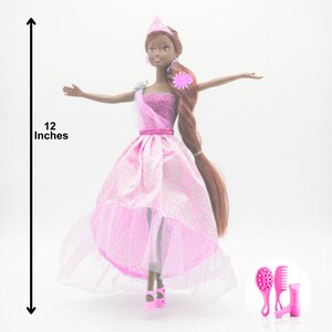 May include: A 12-inch doll in a pink princess gown with a matching tiara and shoes. The doll has long brown hair and is accessorized with pink earrings and a flower. Includes pink hair accessories.