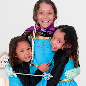 3 girls dress as fairy tale princess, happy and smiling, wearing a blue princess elsa coronation dress and purple cape with a wand, gloves, and princess accessory for kids