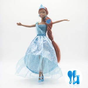 May include: A fashion doll with brown skin, wearing a light blue gown with a high-low hemline and a matching crown. Her long, auburn hair is styled in a braid. Accessories include blue shoes and a hairbrush set.
