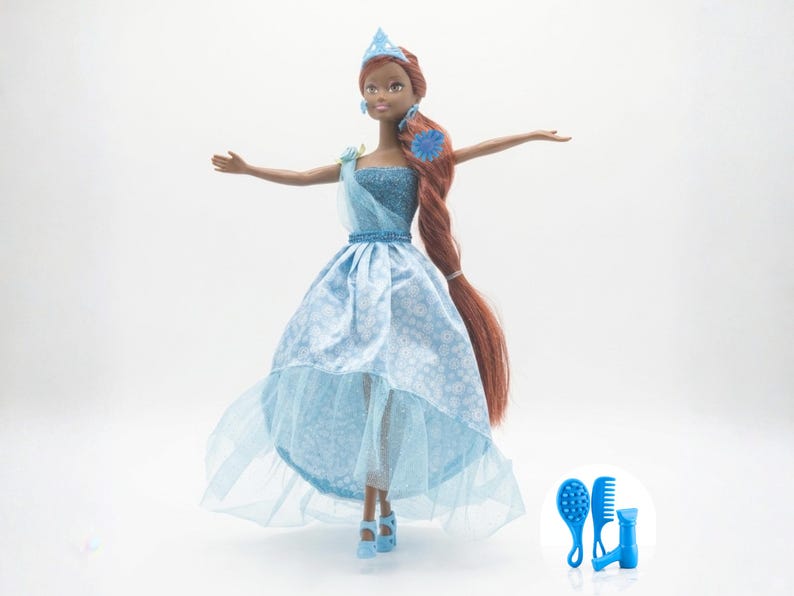 May include: A fashion doll with brown skin, wearing a light blue gown with a high-low hemline and a matching crown. Her long, auburn hair is styled in a braid. Accessories include blue shoes and a hairbrush set.