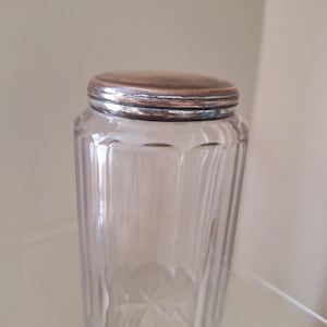 May include: A clear glass jar with vertical fluted sides and a starburst pattern on the base. The container features a circular, metallic lid with a tarnished copper-toned finish and a silver-colored rim, providing a secure top closure.