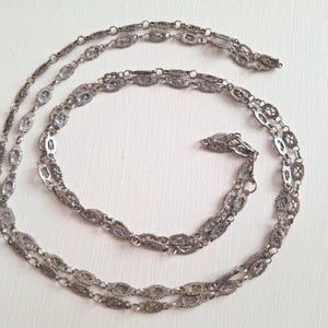 May include: A long silver-toned chain necklace featuring a series of repeating oval-shaped filigree links connected by small circular jump rings. The metal has a darkened, oxidized finish within the intricate wirework patterns of each oval link.