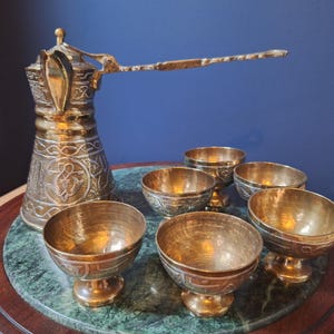 May include: A brass coffee pot with a long handle and an engraved body, accompanied by five matching small brass cups. The pot and cups feature intricate etched patterns throughout their surfaces, showcasing a metallic gold finish and a classic design.