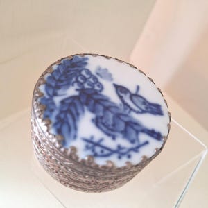 May include: A small round metal trinket box with a decorative textured silver-toned base and a circular lid featuring a white ceramic inlay. The inlay displays a blue painted design of a bird perched on a branch surrounded by foliage.