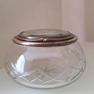 May include: A round glass trinket box featuring a clear, cut-glass base with a geometric diamond pattern. The lid is topped with a circular glass insert framed by a tarnished brass-toned metal rim, creating a vintage-inspired storage container.