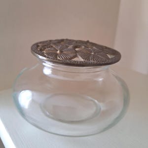 May include: A clear glass jar with a decorative metal lid featuring a floral design. The jar has a rounded shape and sits on a white surface. The lid is a bronze color and adds a vintage touch to the piece.
