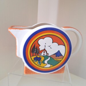May include: A white ceramic creamer with an orange textured rim and handle. The side features a circular graphic depicting a house with a chimney, a winding path, trees, and a mountain under a cloud, framed by concentric blue, orange, and yellow rings.