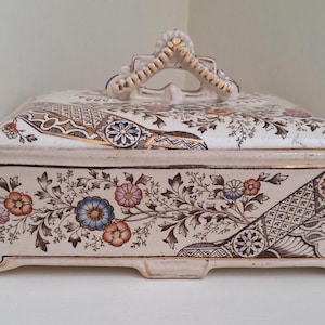 May include: A rectangular ceramic lidded box with a cream-coloured base, featuring intricate floral patterns in blue, orange, and pink, alongside geometric black and gold accents. The lid has a central handle and side handles with gold trim.