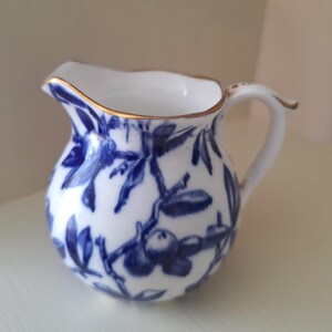 May include: A small white porcelain cream jug featuring a blue botanical pattern of berries and leaves. The pitcher has a curved handle and a gold-trimmed rim, with a rounded body and a distinct pouring spout, showcasing a classic ceramic design.