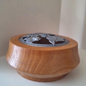 May include: A wooden box with a decorative metal lid. The box is made of light brown wood and has a round shape with a wider base. The lid features a floral design in a silver-colored metal.