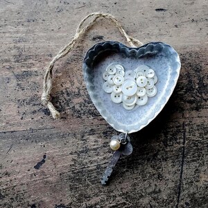 May include: A heart-shaped, silver-colored metal dish filled with white buttons, with a jute string for hanging. A small key with a pearl charm hangs below. The dish sits on a rustic, wooden surface.
