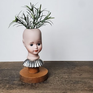 May include: A vintage doll head planter with a light pink face, blue eyes, and red lips. The doll head is mounted on a wooden base with a silver collar and filled with green faux foliage. The planter is a unique decorative item.