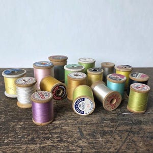 May include: A collection of vintage thread spools in various colors, including yellow, pink, green, and blue. The spools are made of wood and have paper labels with text and designs. The spools are arranged on a dark wooden surface.
