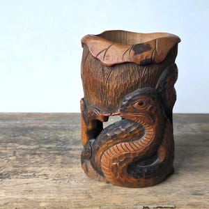 May include: A cylindrical brown wooden brush holder featuring intricate hand-carved details of a coiled snake and a large lotus leaf. The wood displays a natural grain with dark brown highlights and a textured finish throughout the carving.