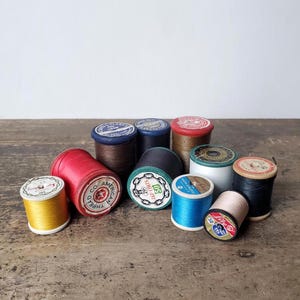 May include: A collection of vintage thread spools in various colors, including red, blue, gold, and black. The spools have labels with text and are arranged on a wooden surface. The spools are of different sizes and shapes.
