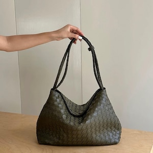 Puede incluir: Un bolso hobo de color verde oliva con una construcción de cuero tejido que presenta un patrón de trenzado distintivo. El bolso tiene una silueta holgada y desestructurada, e incluye dos correas de hombro integradas también tejidas.
