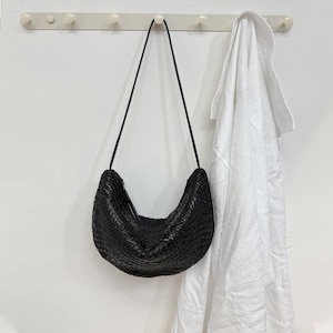 Può includere: Una borsa a tracolla nera intrecciata con una lunga tracolla nera è appesa a una parete bianca. La borsa ha una forma a mezzaluna e una superficie strutturata. Un capo di abbigliamento bianco è appeso a destra della borsa.