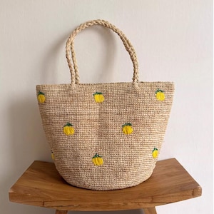 Può includere: Borsa tote in tessuto beige con manico intrecciato. La borsa presenta ricami di frutta gialla. La borsa è appoggiata su una panca di legno. La borsa è di colore marrone chiaro.
