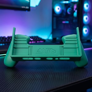 Peut inclure: Une poignée de console de jeu portable sarcelle au design ergonomique avec des poignées profilées et une découpe centrale. La surface avant affiche le texte en relief AYA, avec des fentes et des ouvertures intégrées le long du bord inférieur.