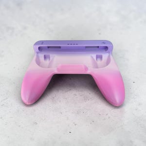 Peut inclure: Une station de charge pour manette avec une finition dégradée lisse passant d'un violet clair en haut à un rose vif à la base. L'appareil présente une zone centrale évidée pour une manette et une fente supérieure avec de petites icônes.