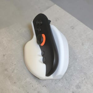 Peut inclure: Une manette de jeu noire avec un accent circulaire orange vif autour du joystick, reposant solidement dans un support blanc incurvé et lisse. La manette présente plusieurs boutons noirs et un design ergonomique maintenu droit par la base.