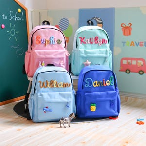 May include: Four backpacks in pink, light blue, mint green, and royal blue. Each bag features a large, colourful, textured name patch on the front pocket: Sophie, Kaitlyn, William, and Daniel. Small decorative patches include a plane and pineapple.