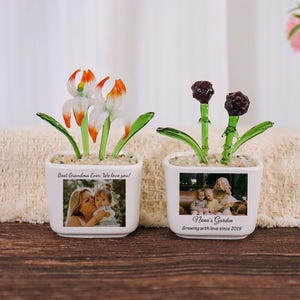 May include: Two white ceramic square planters containing glass flowers. The left planter features a photo of two people and the text Best Grandma Ever. We love you! The right planter features a photo of three people and the text Nana's Garden.