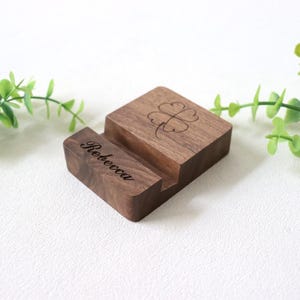 May include: A wooden phone stand with a dark brown finish. The stand features a slot for a phone and a four-leaf clover design. The name "Rebecca" is engraved on the front.