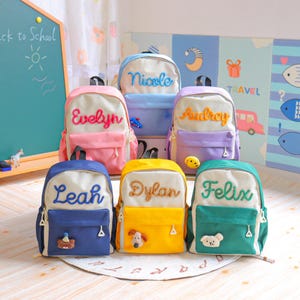 May include: A collection of six colourful backpacks, each featuring a cream-coloured upper section with a personalised name embroidered in cursive, a solid-coloured lower pocket, and a small plush character accent attached to the front zip.