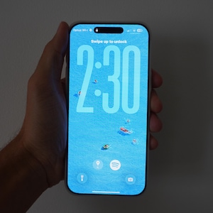 Può includere: Schermo di smartphone che mostra l'ora 2:30 in numeri bianchi su sfondo blu. Lo schermo presenta una scena nautica con piccole barche. Il testo "Swipe up to unlock" è visibile in alto.