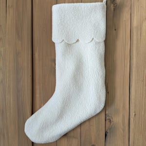 Peut inclure: Une chaussette de Noël blanche avec un bord festonné et une boucle de suspension. La chaussette est faite d'un matériau doux et texturé et est suspendue sur un fond en bois. Une décoration de vacances festive.