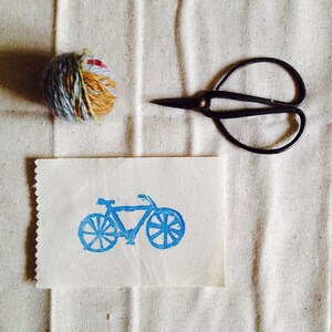 May include: A white card with a blue bicycle print on it. The card is sitting on a white fabric surface with a ball of yarn and a pair of scissors.