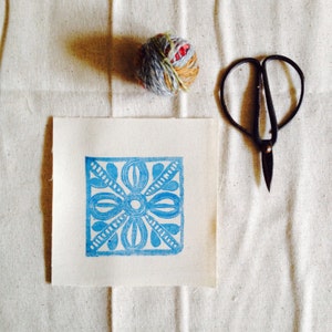 May include: A blue floral design printed on a white fabric square. A ball of yarn and a pair of scissors are also visible on the fabric.