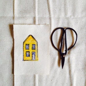 May include: A yellow house with blue windows and a blue door is painted on a piece of white fabric. A pair of black scissors is on the right side of the image.