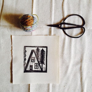 May include: A black and white linocut print of a house with two trees in front of it. The print is on a white piece of paper.