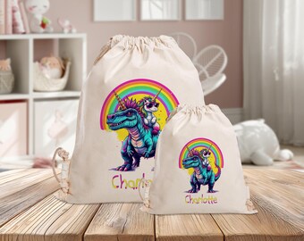 Könnte beinhalten: Zwei beigefarbene Kordelzugbeutel, einer groß und einer klein. Jeder Beutel hat eine farbenfrohe Illustration eines Dinosauriers, der von einem Einhorn unter einem Regenbogen geritten wird. Der große Beutel hat den Namen "Charlotte" in gelber Schrift.