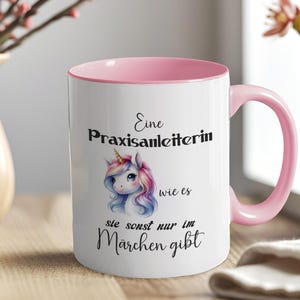 Könnte beinhalten: Weiße Keramiktasse mit rosa Innenraum und Henkel. Die Tasse zeigt eine farbenfrohe Illustration eines Einhorns und Text auf Deutsch. Die Tasse steht auf einer Holzoberfläche.