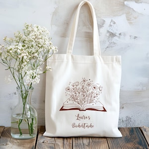 Può includere: Borsa tote color crema con un disegno floreale marrone che esce da un libro aperto. Il testo "Laura's Büchertasche" è stampato sotto l'illustrazione. La borsa ha due manici ed è appoggiata su una superficie di legno.