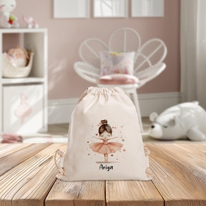 Puede incluir: Una bolsa de cord&oacute;n blanca con una ilustraci&oacute;n de bailarina rosa y el nombre "Ariya" impreso en ella.
