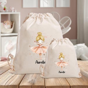 Puede incluir: Dos bolsas de cord&oacute;n beige con una ilustraci&oacute;n de bailarina y el nombre "Aurelie" impreso en cada una. La bailarina lleva un tut&uacute; rosa y tiene el pelo rubio.