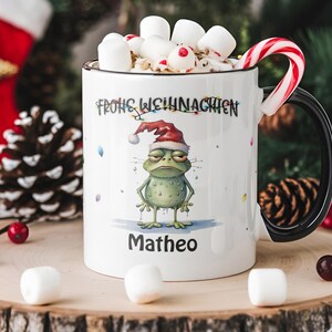 Könnte beinhalten: Weiße Keramik-Tasse mit schwarzem Henkel und Rand, gefüllt mit Marshmallows und einer Zuckerstange. Die Tasse zeigt einen Cartoon-Frosch mit Weihnachtsmütze, dem Text "Frohe Weihnachten" und dem Namen "Matheo".