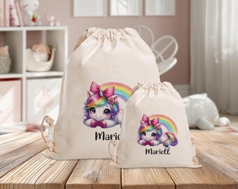 Könnte beinhalten: Zwei weiße Kordelbeutel mit einem Regenbogen-Einhorn-Design. Der größere Beutel trägt den Namen "Marie" aufgedruckt. Der kleinere Beutel trägt den Namen "Mariell" aufgedruckt.