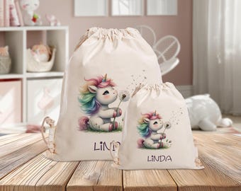Könnte beinhalten: Zwei beigefarbene Kordelzugbeutel mit einem bunten Einhorn-Design. Das Einhorn hat eine Regenbogenmähne und einen Regenbogen-Schweif und hält einen Löwenzahn. Der Name "LINDA" ist auf der größeren Tasche und "LINDA" auf der kleineren Tasche aufgedruckt.