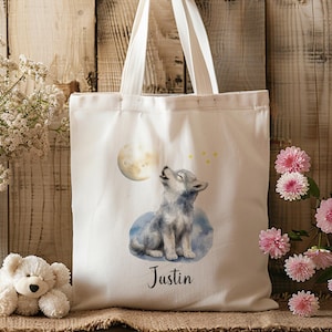 Könnte beinhalten: Eine weiße Stofftasche mit einer Aquarellillustration eines grauen Wolfswelpen, der einen gelben Mond anheult, begleitet von kleinen gelben Sternen. Unter dem Wolf ist der Name Justin in einer schwarzen Schreibschrift aufgedruckt. Zwei weiße Henkel.