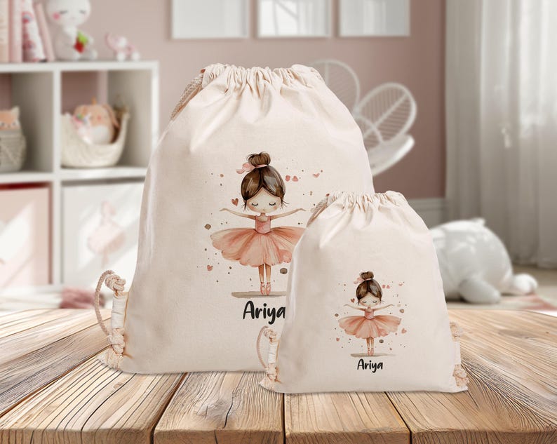 Puede incluir: Dos bolsas de cord&oacute;n beige con una ilustraci&oacute;n de bailarina y el nombre "Ariya" impreso en ellas. La bailarina lleva un tut&uacute; rosa y tiene el pelo casta&ntilde;o.
